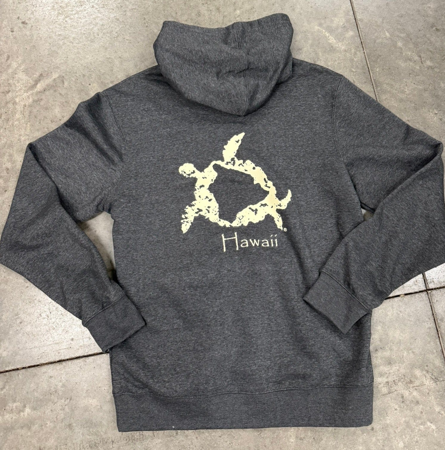 Gray hoodie with a turtle design and 'Hawaii' text on the back, laid flat on a concrete surface.