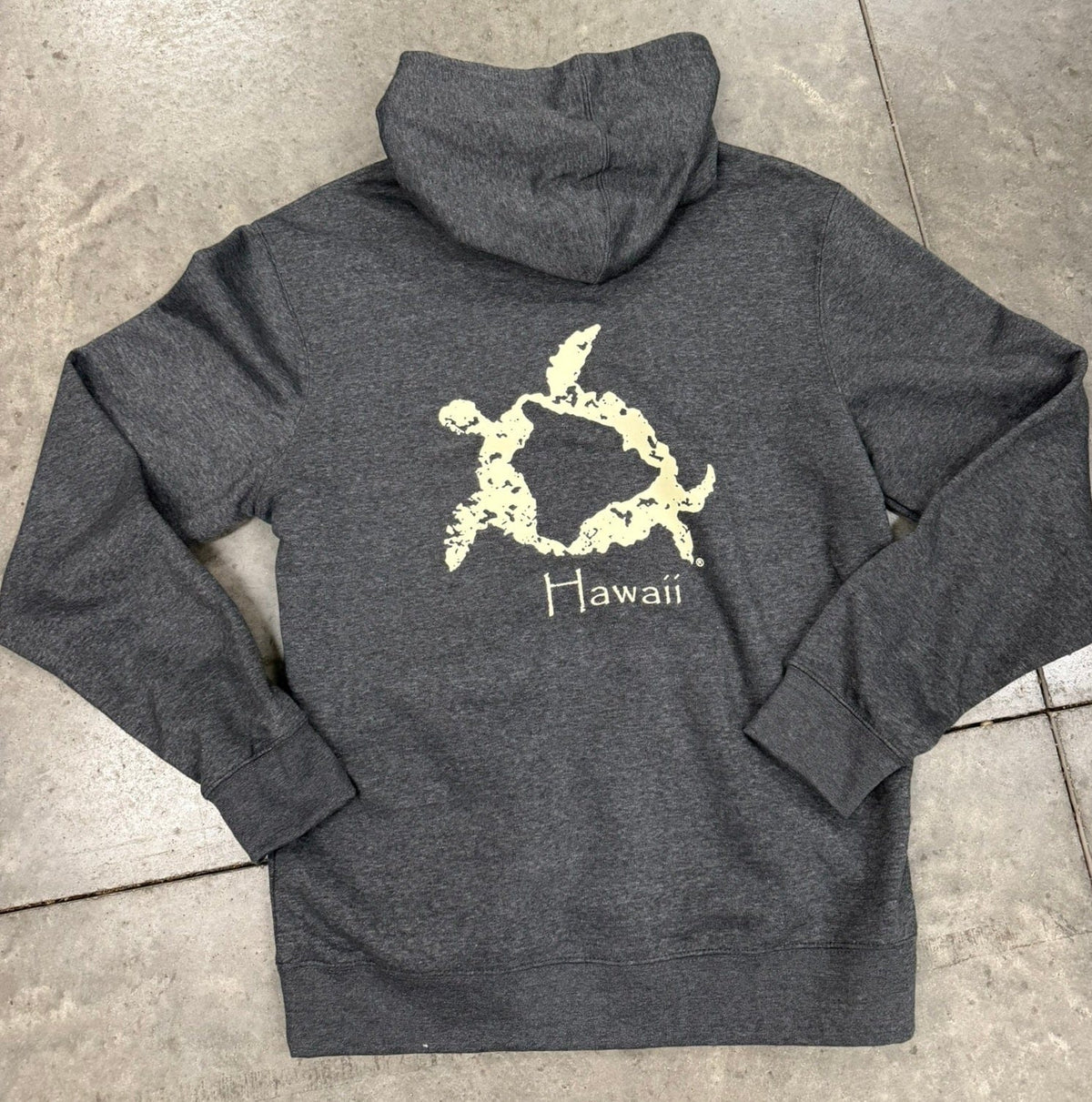 Gray hoodie with a turtle design and &#39;Hawaii&#39; text on the back, laid flat on a concrete surface.